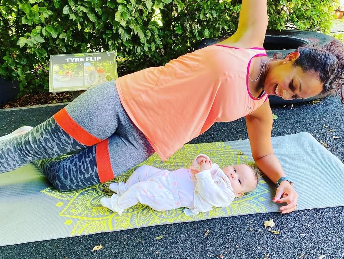 A mum holding a plank on a mat outdoors with her baby lying alongside her