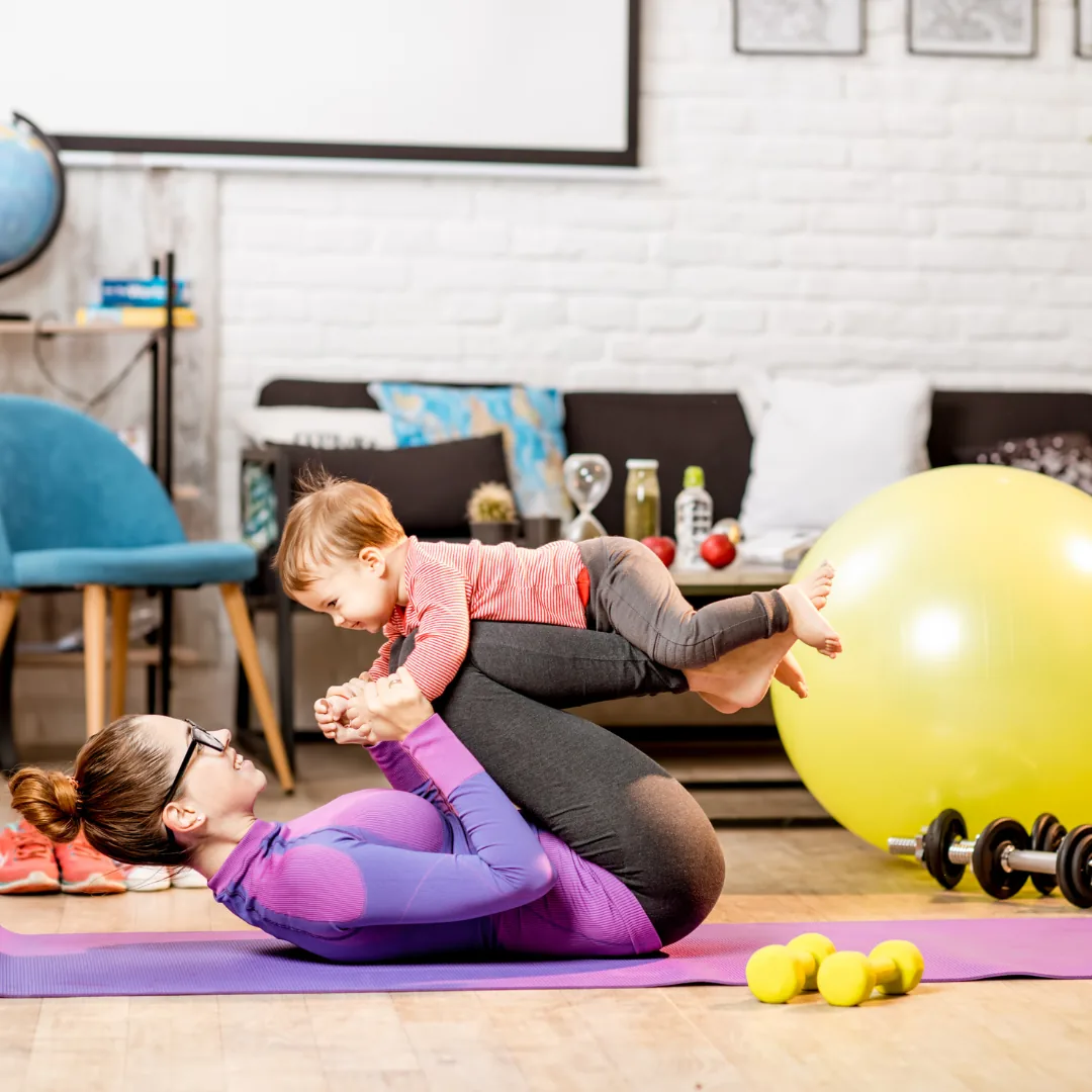 A mother lying on an exercise mat lifting her smiling toddler above her during a home workout